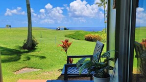 A patio with a bench and potted plants, overlooking a grassy area with a view of the ocean and palm trees. - Princeville corner unit w/AC & pool for up to 6*; Ocean/Sunset/Golf/Mtn Views (Princeville)