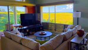 A spacious living room with a large flat-screen TV, comfortable seating, and a view of a green field through large windows. - Princeville corner unit w/AC & pool for up to 6*; Ocean/Sunset/Golf/Mtn Views (Princeville)