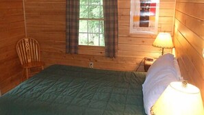 Una habitación de cabaña de madera con una cama, una silla, una ventana con cortinas, una lámpara y un cartel en la pared.