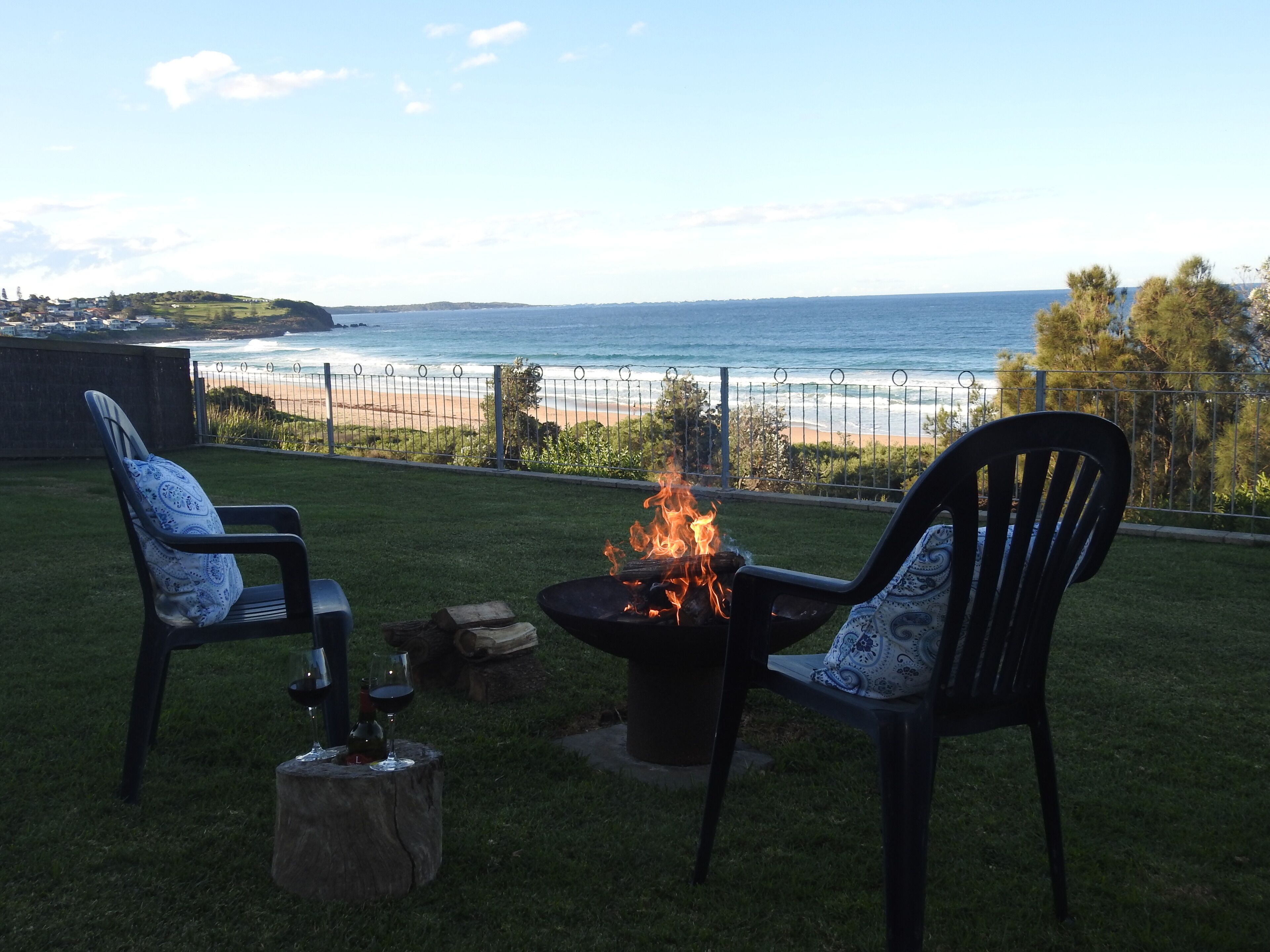 Une terrasse avec deux chaises, un foyer extérieur et un verre de vin. On aperçoit une plage et l’océan au loin.