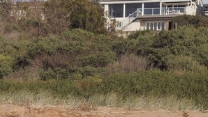 Una casa frente a la playa con balcón, rodeada de vegetación exuberante y un primer plano arenoso.
