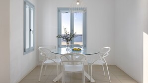 A dining area with a glass table, white chairs, a vase with flowers, and a view of the sea through large windows. - Private pool, Breathtaking Panoramic View to the Sea & Sunset (Paros)