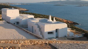A coastal building with white walls and a stone foundation, overlooking a calm sea with distant mountains. - Private pool, Breathtaking Panoramic View to the Sea & Sunset (Paros)