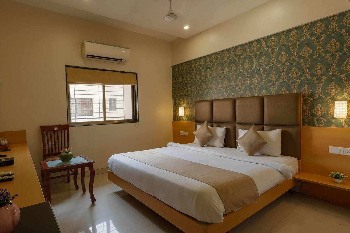 A hotel room with a bed, a TV, a desk with a vase, a chair, and a window.