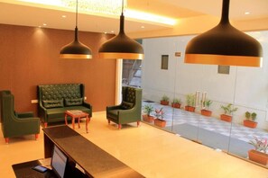 A reception area with a reception desk, two green armchairs, a small table, and a large window with potted plants. - Magnus Square Business Hotel (Pune)