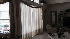 A living room with a sofa, a coffee table, and a TV. There is a window with curtains and a view of the outside.