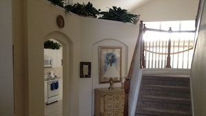 A hallway leading to a kitchen with white cabinets and a microwave, a wooden dresser, and a staircase with a wooden railing.