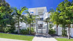 A modern multi-story building with a central entrance, surrounded by lush greenery and palm trees.
