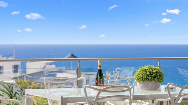 A table with a bottle of champagne, a glass, and a potted plant on a balcony overlooking the sea.