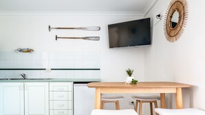 A modern kitchen with white cabinets, a wooden table, two white bar stools, a flat-screen TV, and a decorative mirror. - Surfrider | Bright Beachfront Apartment | Mollymook Beach (Mollymook Beach)