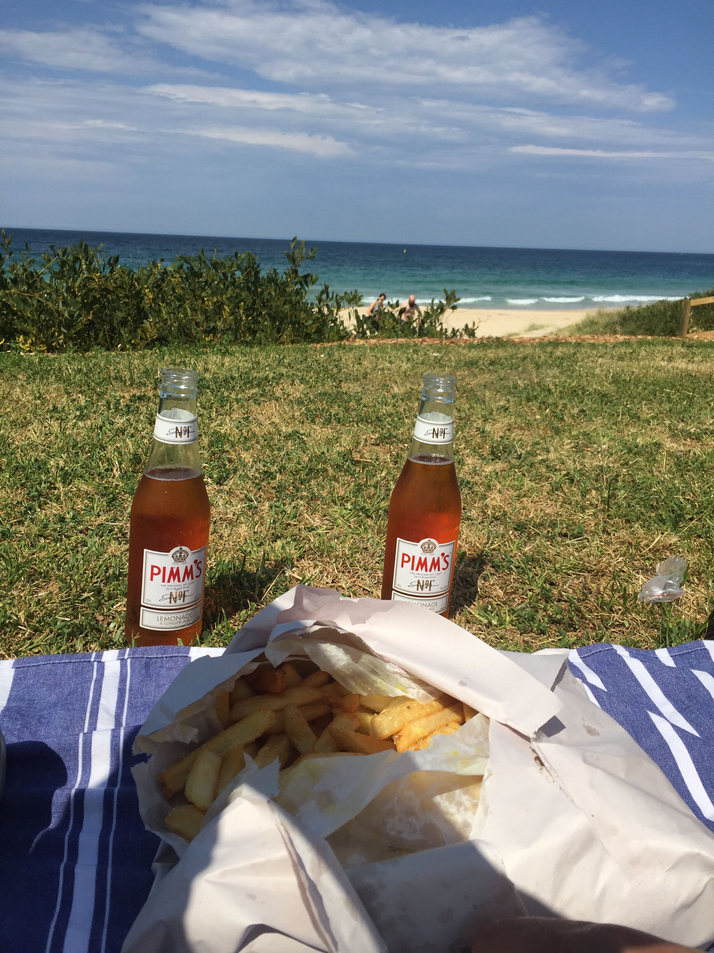 A picnic setup with two bottles of Pimm's No. 1 lemonade, a basket of fries, and a blanket on a grassy area near the beach.