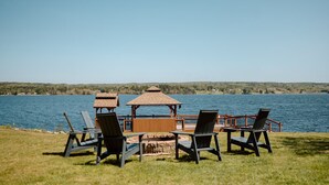 A lakeside outdoor seating area with wooden tables and chairs, a gazebo, and a view of the water and distant hills. - The Cedar- Waterfront /SAUNA /Kayaks /Swimming (Cape Breton Regional Municipality)