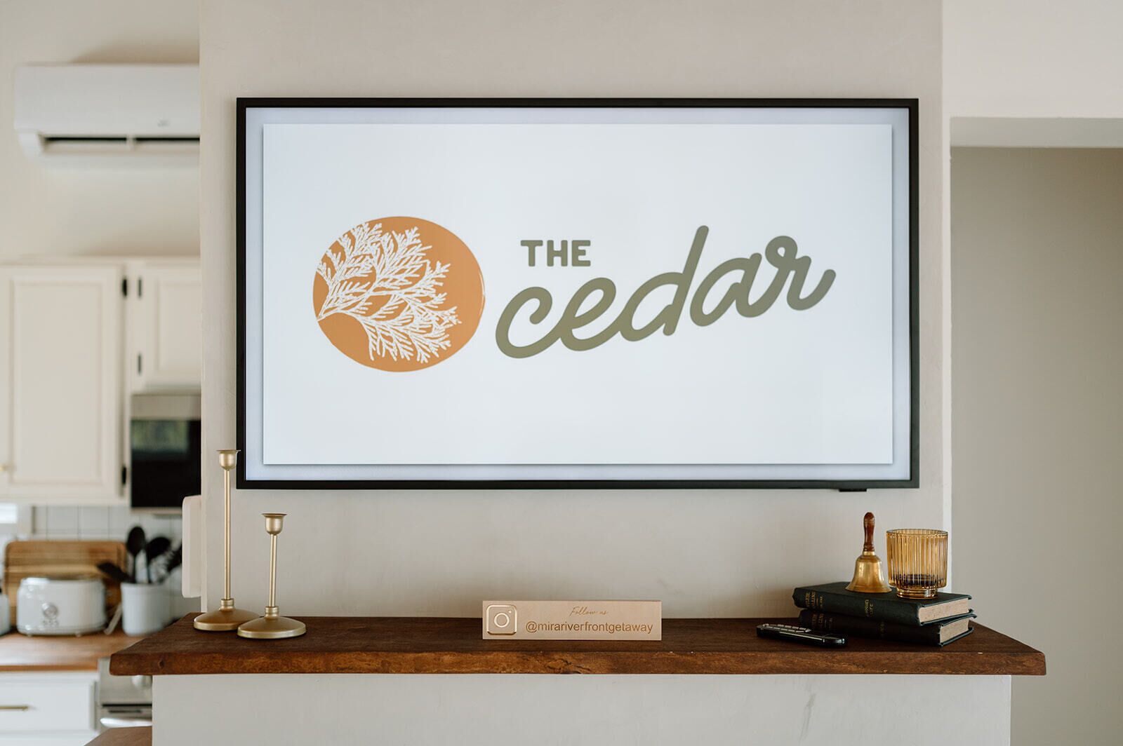 A framed sign with the text "THE CEDAR" and a tree logo, placed on a wooden counter with a nameplate, books, and a remote control.