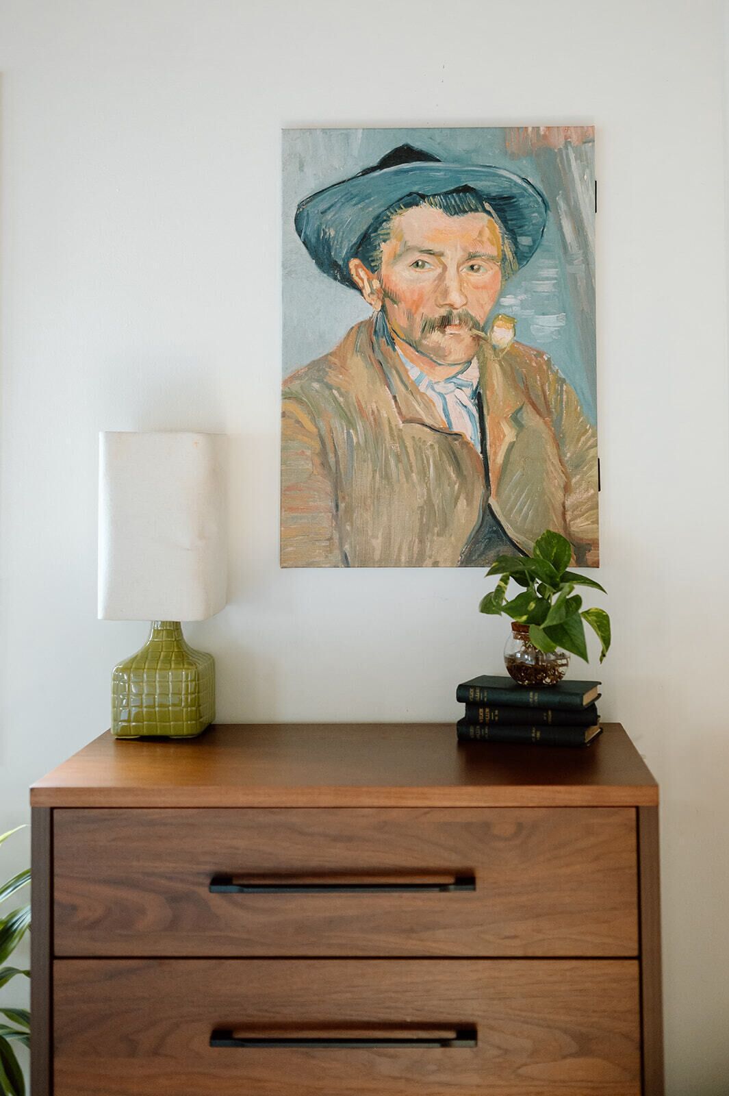 Un tableau représentant un homme portant un chapeau, accroché à un mur, avec une lampe, une plante et une pile de livres posés sur un meuble en bois.