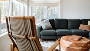 A modern living room with a dark grey sofa, a wooden chair, and a wooden coffee table with a book. - The Cedar- Waterfront /SAUNA /Kayaks /Swimming (Cape Breton Regional Municipality)