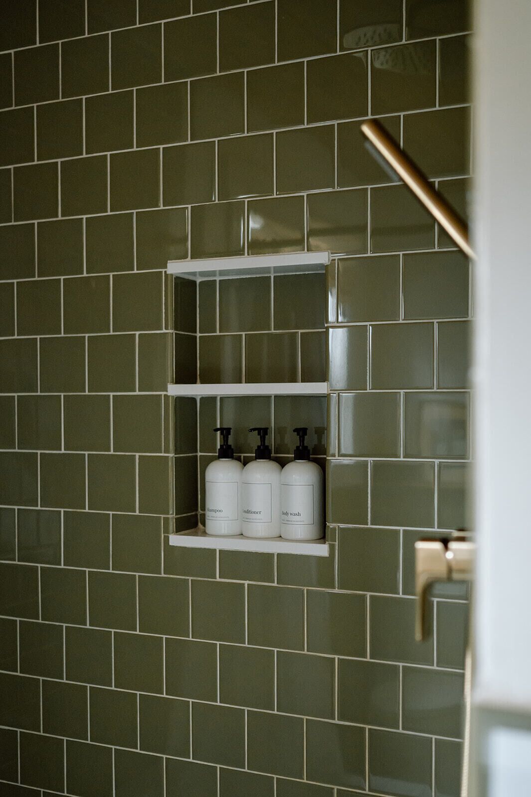 A bathroom with green tiles, a shelf with three bottles of shampoo, conditioner, and body wash.
