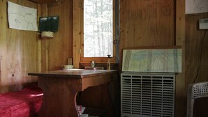 Interior de una cabaña de madera con un escritorio de madera, una ventana con vistas a árboles, un aire acondicionado de pared y una colcha roja.