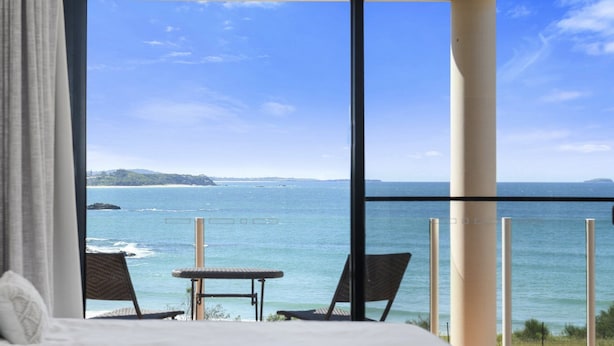 A bedroom with a large window overlooking a beach and ocean. There is a bed, two chairs, and a small table.