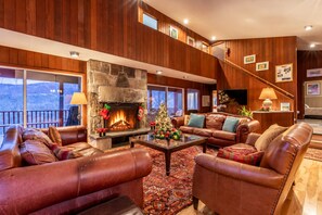A cozy living room with leather sofas, a stone fireplace, a Christmas tree, and a staircase leading to an upper level.