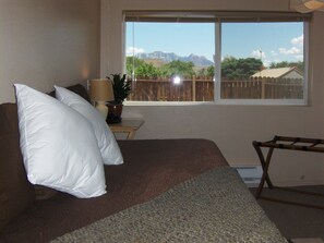 A bedroom with a bed, pillows, a nightstand with a lamp and a plant, a window with a view of mountains and a fence. - Zion National Park Views House on Private Lot. No Cleaning Fee! (Virgin)
