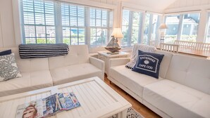 A bright, sunlit living room with white furniture, large windows, and a view of the outside. - New Seabury Beach Cottage with stunning ocean views, steps to private beach (Mashpee)