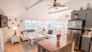 A spacious living area with a white sofa, coffee table, and a kitchen with a refrigerator and cabinets. - New Seabury Beach Cottage with stunning ocean views, steps to private beach (Mashpee)