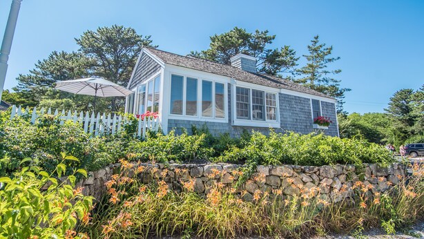 A house with a stone wall, a white picket fence, and a clear blue sky. - New Seabury Beach Cottage with stunning ocean views, steps to private beach (Mashpee)
