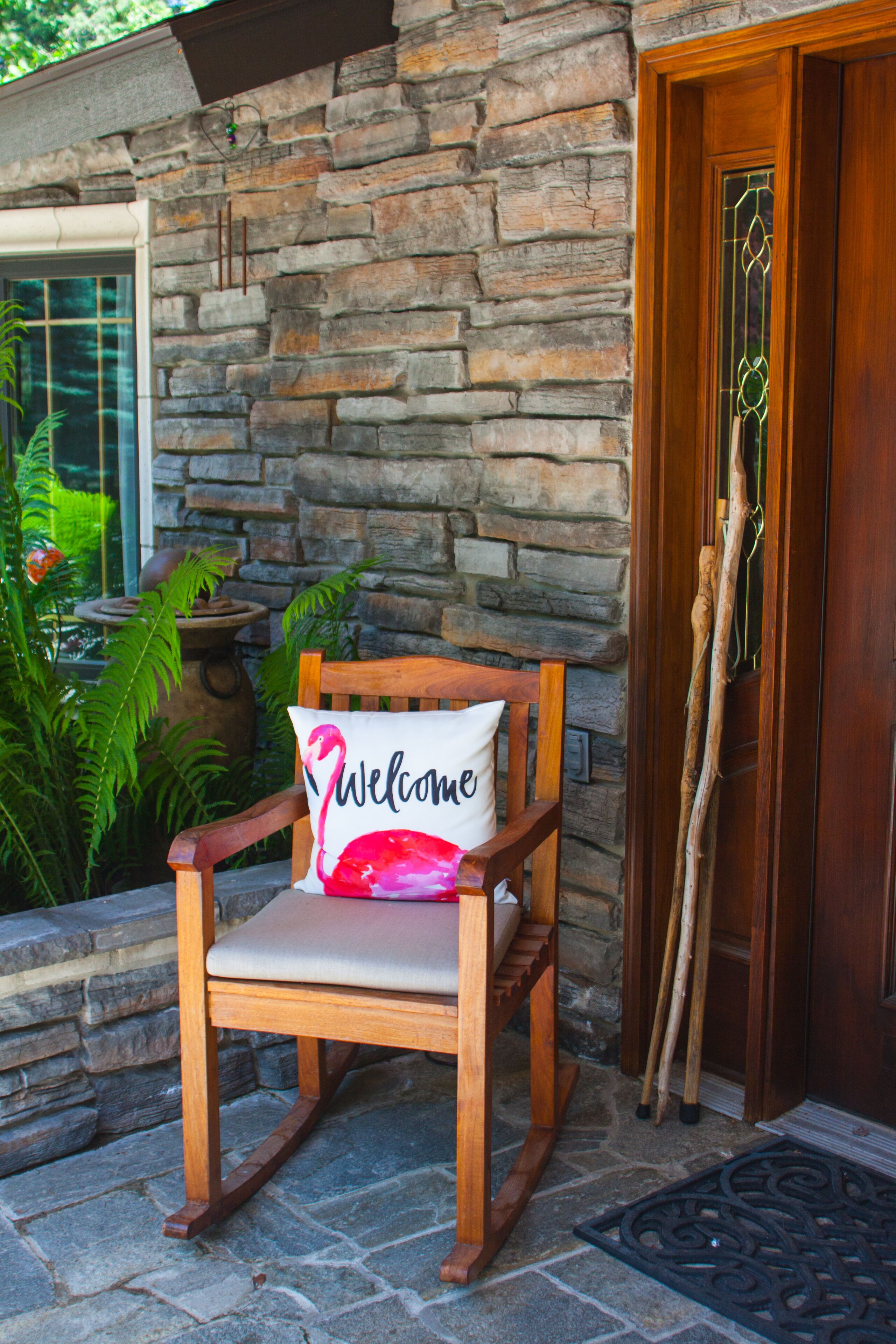 A wooden rocking chair with a 'Welcome' pillow, positioned outside a stone building with a wooden door.