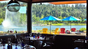 Restaurante con vistas a un lago y bosque, mesas al aire libre con sombrillas y una lámpara colgante.
