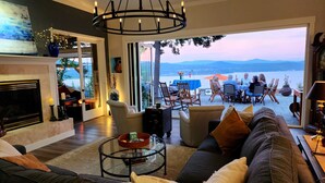Amplia sala de estar con un sofá grande, una mesa de centro de vidrio y una chimenea. Hay un comedor con asientos exteriores y vistas al agua.