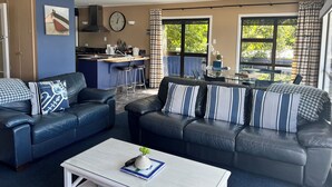 A modern living room with two black leather sofas, a coffee table, and a kitchen area with a clock and bar stools. - Beachfront Bliss ~ Upper Deck Apartment (Coromandel Township)