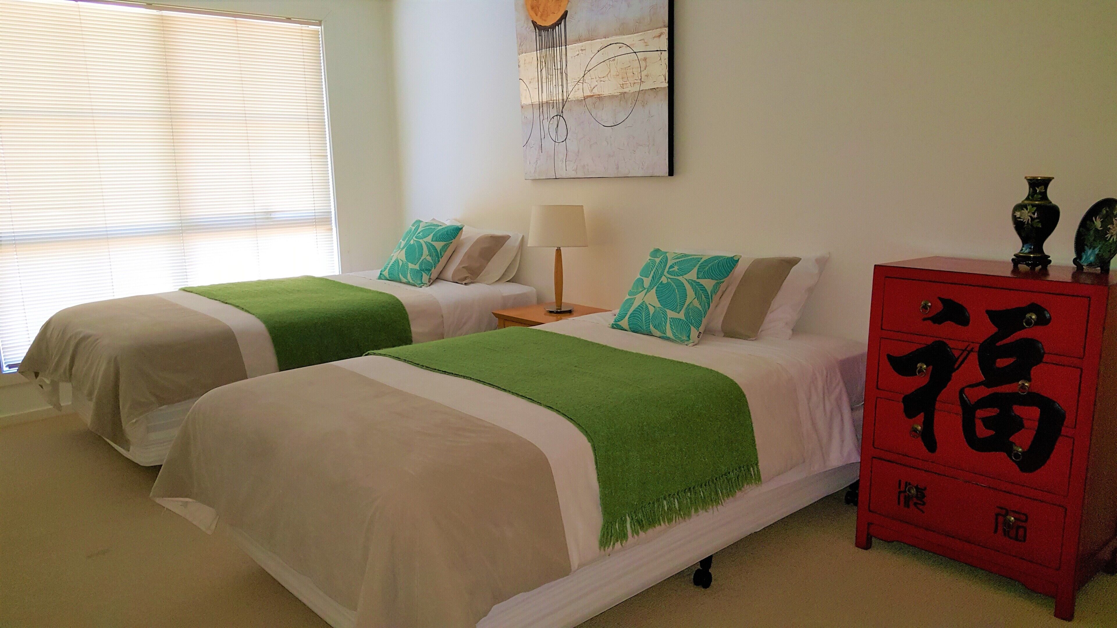 A hotel room with two beds, each with a green throw blanket, white bedding, and matching pillows. There is a red cabinet with Chinese characters, a painting on the wall, and a window with blinds.