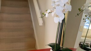 Eine Treppe mit beigem Teppich und weißen Wänden, ein roter Kommode mit einer weißen Orchidee und ein Spiegel, der die Orchidee reflektiert.