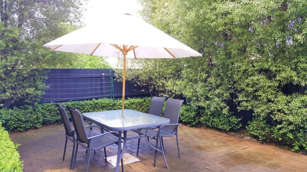 An outdoor patio with a table and chairs, a large umbrella, and a hedge in the background.