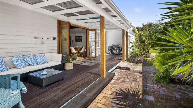 A covered outdoor seating area with a wooden deck, a white wall, and a garden with tropical plants.