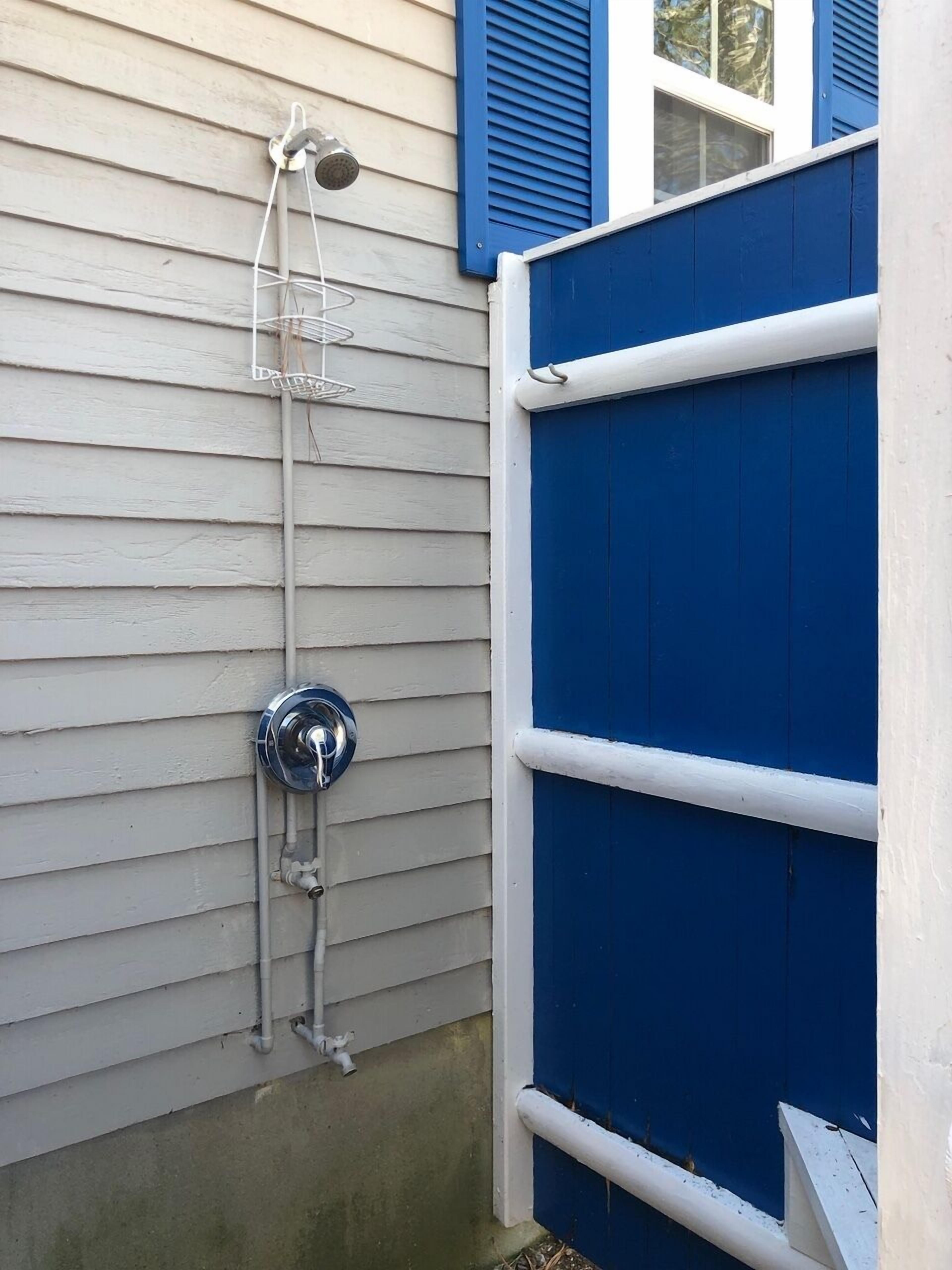 A small, enclosed outdoor shower area with a metal showerhead and hose, attached to a white exterior wall.