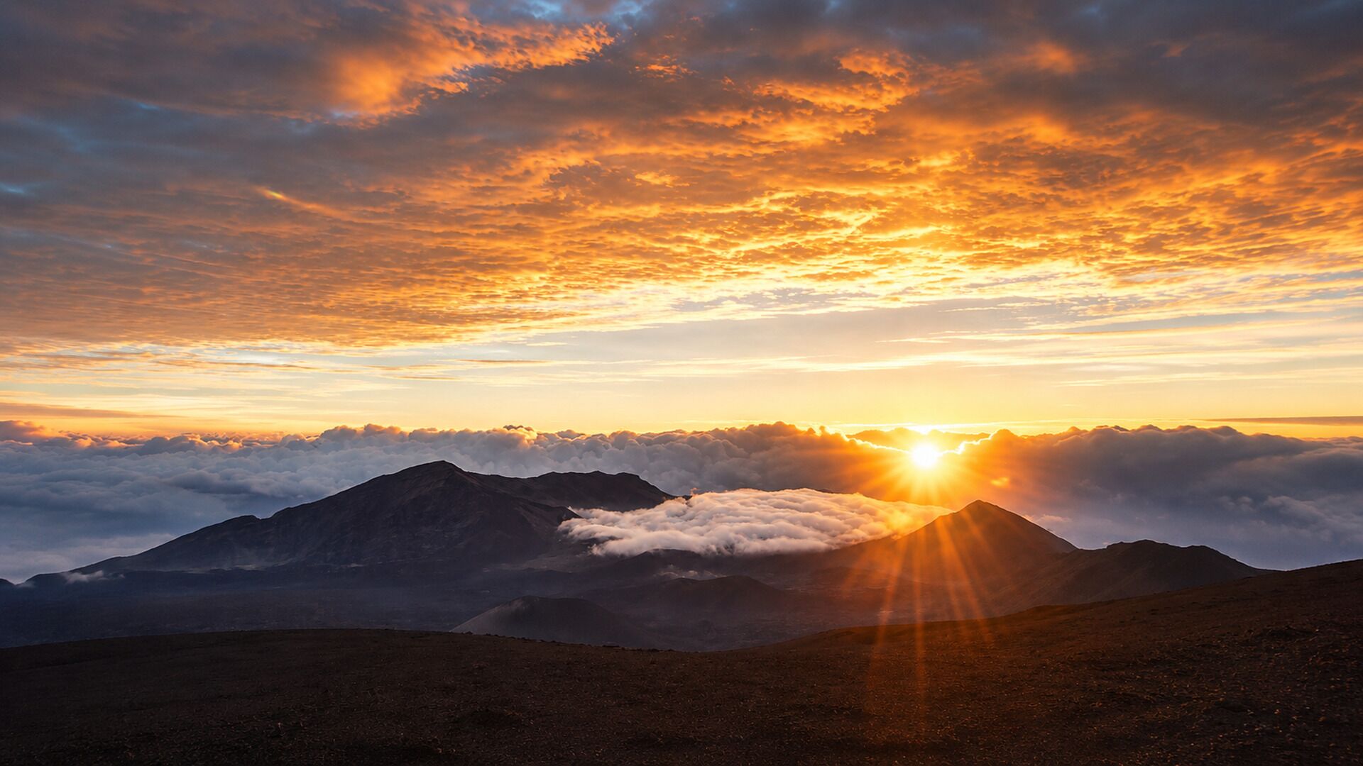 A mountain landscape with a sunrise or sunset, clouds below the peaks, and no visible structures or people.