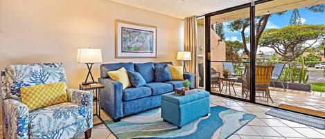 A living room with a blue sofa, a patterned armchair, and a coffee table. There is a ceiling fan, a framed picture on the wall, and a view of the outdoors through a glass door.