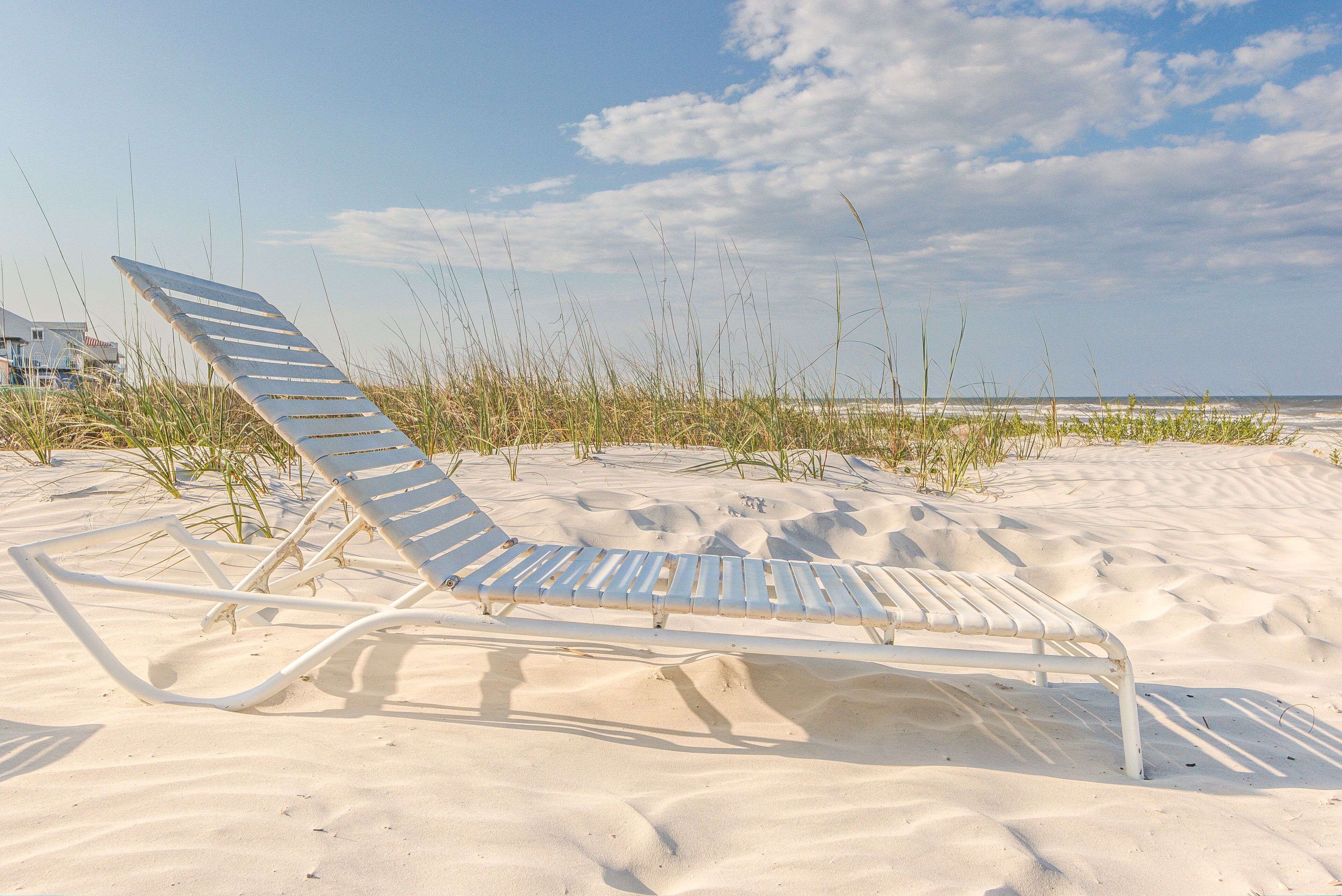 A beach chair on a sandy beach with grass and a clear sky in the background.