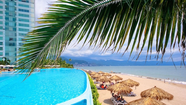 A swimming pool adjacent to a beach with thatched umbrellas and lounge chairs.