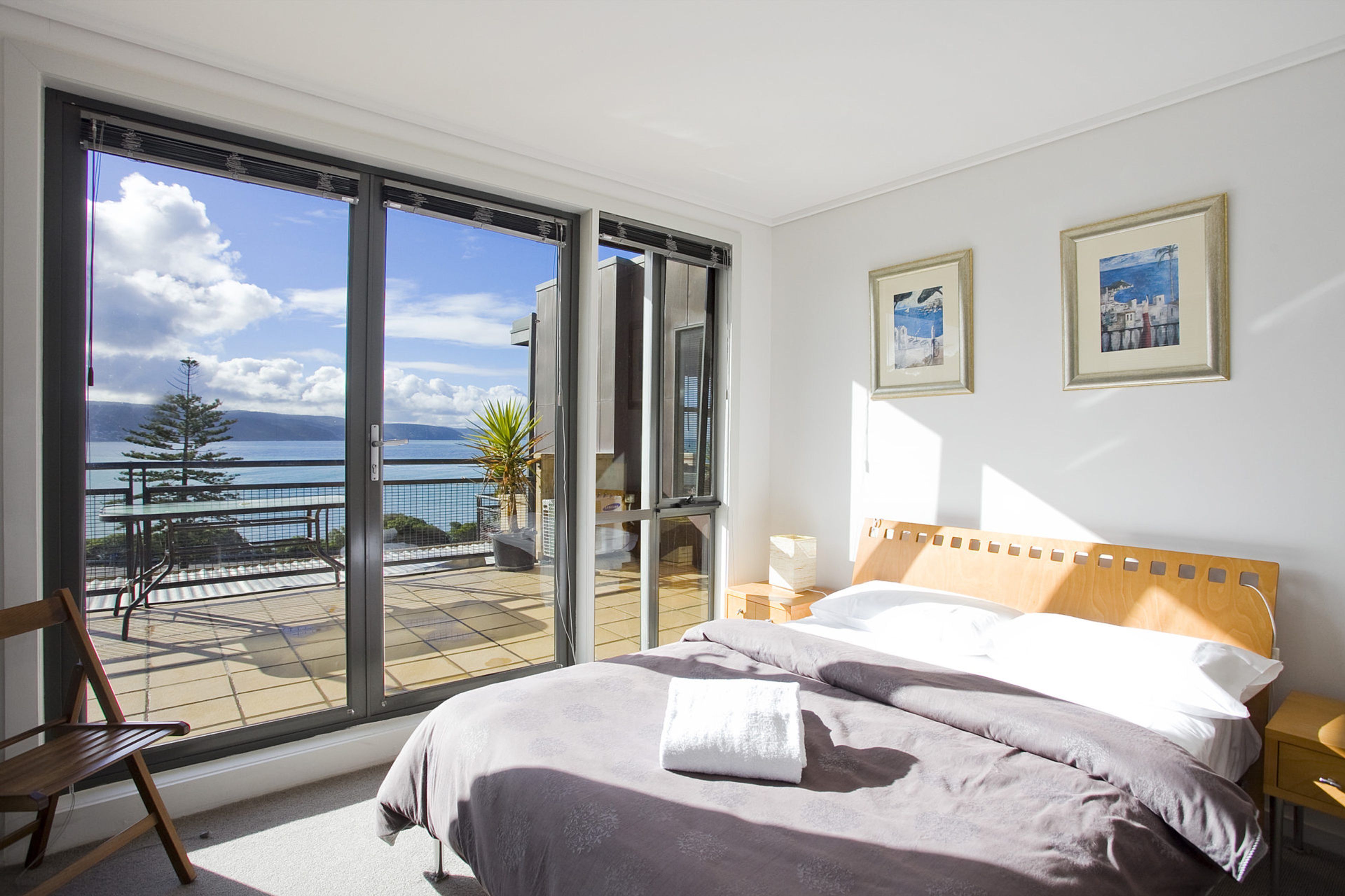 A bedroom with a large bed, a view of the sea through floor-to-ceiling windows, and two framed pictures on the wall.