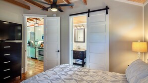 Un dormitorio con cama, televisor, ventilador de techo y puerta corredera que da a una cocina.