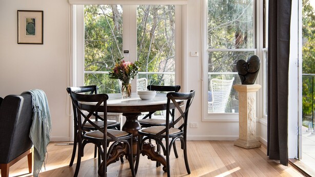 A dining area with a round table, four chairs, a vase with flowers, and a statue on a pedestal. - Lake Como Villa- Lake Daylesford on your doorstep (Daylesford)