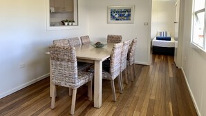 A dining area with a wooden table and wicker chairs, a kitchen area with cabinets, and a bed with a window. - 35 YAMBA STREET Dog friendly (Yamba)
