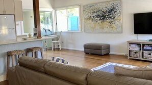 A modern living room with a large beige sectional sofa, a dining area with a white table and chairs, a grey ottoman, a ceiling fan, and a large abstract painting on the wall. - 35 YAMBA STREET Dog friendly (Yamba)