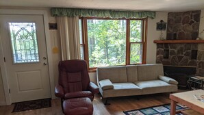 A living room with a stone fireplace, a sofa, a recliner, and a window with a view of trees.