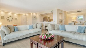 A living room with two white sofas, a coffee table with a floral arrangement, and a kitchen in the background.