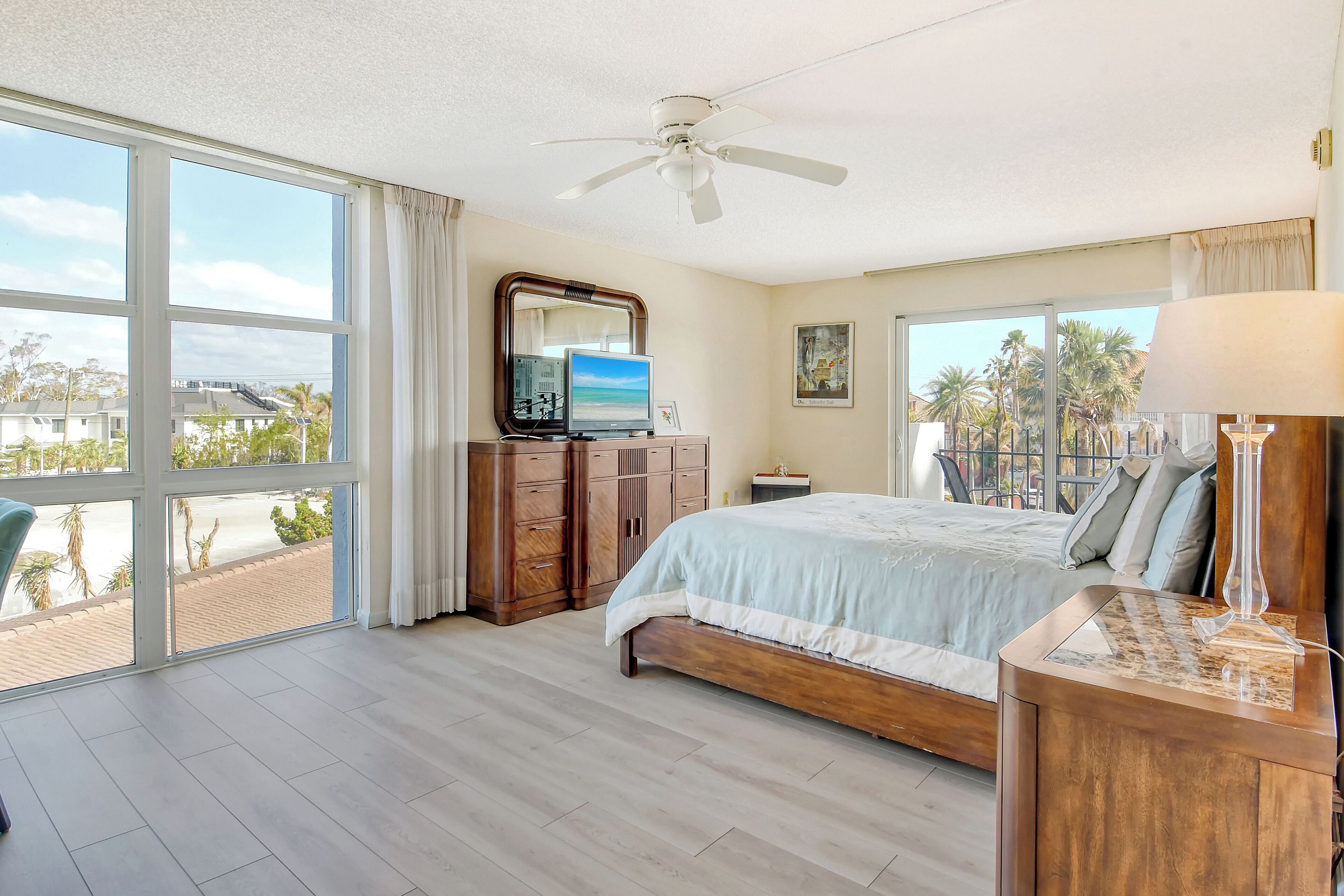 A bedroom with a large bed, a ceiling fan, a TV, and a view of palm trees and buildings through large windows.