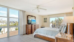 A bedroom with a large bed, a ceiling fan, a TV, and a view of palm trees and buildings through large windows.