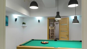 Una mesa de billar con superficie de fieltro verde, lámparas colgantes negras y una puerta de madera al fondo.